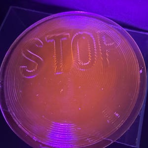 May include: A red, round, plastic stop sign with the word "STOP" embossed on the surface. The sign is illuminated with a purple light.