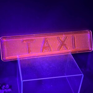 May include: A rectangular, orange-tinted plastic sign with the word "TAXI" in bold, raised letters. The sign has a textured surface and is illuminated against a dark purple background.