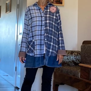 Puede incluir: Una mujer con una camisa a cuadros azul y blanco, una falda vaquera y leggings negros. Una flor rosa está prendida en la camisa. Lleva sandalias negras. El fondo incluye un pasillo con obras de arte y una silla de madera.