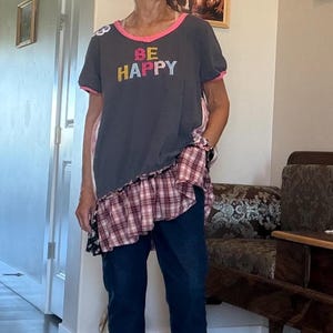 May include: A woman wearing a gray t-shirt with the words "BE HAPPY" in colorful letters. The shirt has pink trim and is paired with dark blue jeans and sandals. A plaid and floral patterned fabric is attached to the shirt.