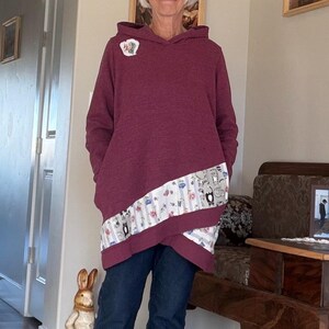 Peut inclure: Un sweat à capuche bordeaux avec un panneau diagonal à motifs de chats. Le sweat-shirt a une poche kangourou et est associé à un jean bleu foncé. Une petite épingle est attachée à la capuche.