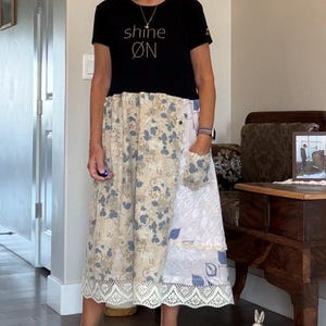 May include: A black short-sleeved t-shirt with the text shine ØN in white lettering, attached to a midi-length skirt featuring a patchwork of beige and blue floral patterns, a side pocket, and a white lace trim along the bottom hem.
