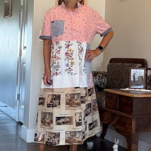 May include: A midi-length dress featuring a pink short-sleeved bodice with a small blue and white gingham pocket. The midsection has a white floral print panel, while the skirt displays a beige pattern of vintage cars and text.