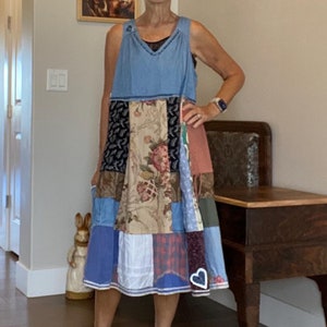 Puede incluir: Una mujer con un vestido de patchwork azul y multicolor con un parche en forma de corazón. Está de pie sobre una alfombra estampada en una habitación con una mesa de madera y obras de arte en las paredes.