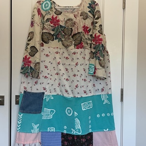 Può includere: Un vestito patchwork con un top a stampa floreale e a foglie, una sezione centrale floreale bianca e una sezione inferiore turchese con disegni di pesci e geometrici. L'abito ha una tasca in denim e un orlo in pizzo rosa.