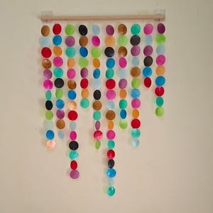 Puede incluir: Adorno de pared colorido hecho de objetos planos y redondos de varios colores. Dos sillas de madera con cojines estampados están debajo, bajo un cuadro enmarcado.