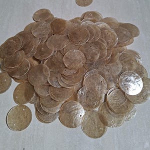 Könnte beinhalten: Eine große Ansammlung von kreisförmigen, durchscheinenden, hellbraunen Muschelscheiben. Jede flache Scheibe hat ein kleines, mittiges Loch zum Auffädeln. Die Scheiben haben eine natürliche, schillernde Textur und liegen in einem losen Haufen, wobei einige in einem Plastikbeutel verbleiben.