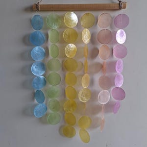 May include: A wall hanging featuring six vertical strands of circular, translucent capiz shells in a gradient of blue, green, yellow, orange, pink, and purple. The strands are suspended from a horizontal wooden dowel with a jute twine hanger.