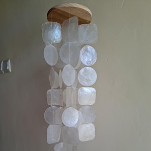Puede incluir: Un carillón de viento decorativo con una parte superior de madera y múltiples hilos de piezas de concha blanca iridiscente en varias formas. Las formas incluyen cuadrados, círculos y formas irregulares, creando un efecto brillante.