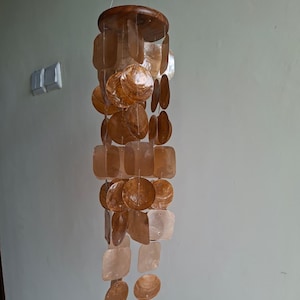 May include: A hanging wind chime featuring a wooden top and multiple shell pieces in diverse shapes and sizes. The shells are mainly brown and tan, with some translucent areas. The chime is suspended by a thin string.