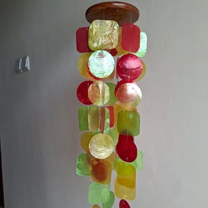 Puede incluir: Un carillón de viento colorido con una parte superior de madera y una cascada de piezas iridiscentes de varias formas en rojo, amarillo y verde. Las formas incluyen cuadrados, círculos y diseños en forma de hoja, creando un adorno colgante decorativo.
