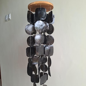 May include: A decorative wind chime featuring a wooden top and a cascade of dark grey shell pieces in various shapes. The shells are suspended by strings, creating a visually appealing and potentially musical ornament. The chime hangs against a plain wall.
