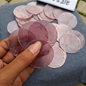 Puede incluir: Una colección de lentejuelas redondas rosas translúcidas con pequeños agujeros, sostenidas en una mano. Las lentejuelas varían en tonalidad de rosa claro a oscuro. Un trozo de papel con el texto "TM sale" está en el fondo.