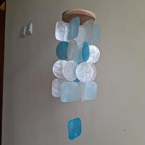 May include: A hanging mobile featuring a circular wooden top from which multiple strands of translucent, iridescent shell pieces are suspended. The shells are shaped as squares and circles in shades of white, pale blue, and bright turquoise.