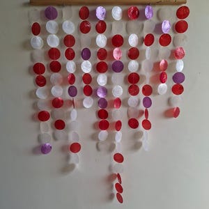 May include: A wall hanging featuring multiple vertical strands of circular, translucent shell discs in shades of red, white, and purple. The strands are suspended from a horizontal wooden rod, creating a cascading, layered arrangement of colourful, flat, round elements.