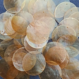 Könnte beinhalten: Eine Ansammlung transluzenter, schillernder, runder Muschelscheiben mit kleinen Löchern am Rand. Die Scheiben variieren in der Farbe von blasscreme bis warmbraun und liegen auf einem blauen Stoff.