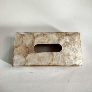 May include: Rectangular tissue box cover with a natural, pearlescent shell mosaic design. The cover has a large oval opening for tissue dispensing. The shell pieces are in shades of cream, beige, and brown.