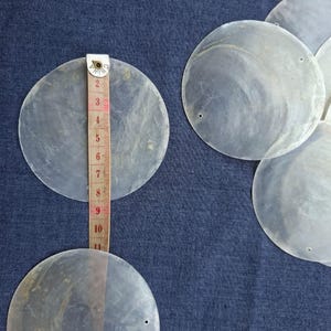Può includere: Diversi dischi rotondi traslucidi di madreperla sono esposti su un tessuto blu. Un metro a nastro mostra che un disco misura circa 10 cm di diametro. I dischi hanno piccoli fori vicino al bordo, suggerendo un uso artigianale o di gioielleria.