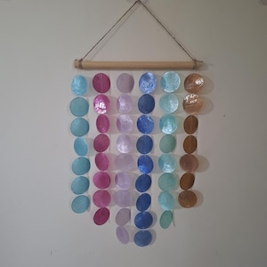 May include: A wall hanging featuring six vertical strands of circular, iridescent capiz shell discs in shades of light blue, magenta, lavender, dark blue, teal, and brown, suspended from a horizontal wooden dowel with a jute twine hanger.