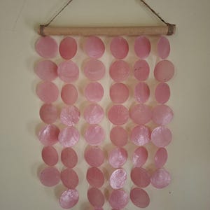 May include: A pink shell wind chime hanging from a wooden dowel and twine. The chime is made of numerous, round, pink shells, each with a slightly iridescent sheen. The shells are strung together in vertical rows, creating a decorative, coastal-themed wall hanging.
