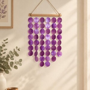 May include: A wall hanging featuring six vertical strands of circular purple capiz shell discs suspended from a horizontal wooden rod. The strands are arranged in a V-shape, with the centre strands being the longest, all attached to a jute rope.