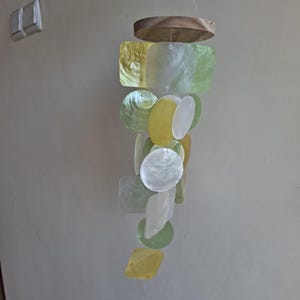 May include: A hanging mobile featuring a circular wooden top piece with multiple strands of translucent, iridescent shell pieces in shades of white, pale yellow, and light green. The shell pieces are shaped as circles, squares, and diamonds.