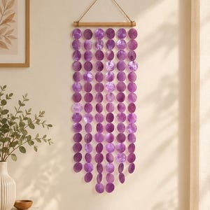 May include: A wall hanging featuring multiple vertical strands of circular, iridescent purple shell discs suspended from a horizontal wooden rod. The strands are arranged in a cascading pattern, with the centre strands longer than the outer edges.