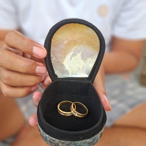 May include: Two gold wedding rings with a small diamond accent are displayed in a black velvet ring box. The box has a decorative, iridescent shell-like interior and is held open, showcasing the rings.