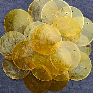 Puede incluir: Un conjunto de discos de concha redondos, dorados e iridiscentes, con pequeños agujeros, dispuestos sobre una tela azul. Las conchas son translúcidas, revelando patrones y texturas sutiles. Cada disco mide unos 5 cm de diámetro.