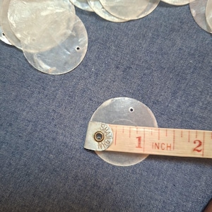 May include: A collection of circular, translucent white mother-of-pearl shell discs, each featuring two small pre-drilled holes. A measuring tape placed across one disc shows a diameter of approximately 3.8 cm for these craft components.