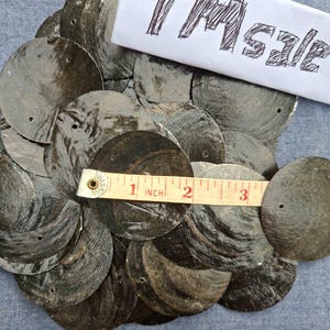 May include: A collection of dark grey, circular, shell-like objects, each with a small hole. A measuring tape shows the objects are approximately 7.6 cm in diameter. The text "TM sale" is written on a piece of paper.