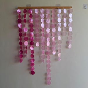 May include: A wall hanging featuring multiple vertical strands of circular pink capiz shells suspended from a horizontal wooden rod. The shells transition in colour from deep magenta on the left to a pale, translucent light pink on the right.