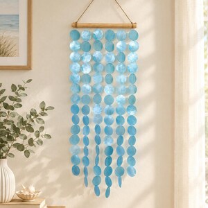 Peut inclure: Une décoration murale composée de multiples brins verticaux de disques en nacre bleu clair irisé, suspendus à une tige en bois horizontale. Les brins sont fixés à la tige par une fine cordelette pour la suspension murale.