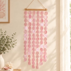 May include: A wall hanging featuring multiple vertical strands of circular, translucent pink shell discs suspended from a horizontal wooden rod. The strands are attached to the rod by a thin twine cord, creating a cascading, layered display.