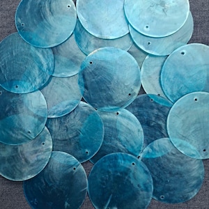 Puede incluir: Colección de discos redondos translúcidos de color verde azulado, con un brillo nacarado. Cada disco tiene un pequeño agujero cerca de la parte superior. Los discos están dispuestos sobre una superficie gris, creando un efecto en capas. Ideal para manualidades o joyería.