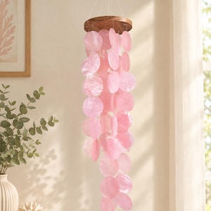 Peut inclure: Un mobile suspendu composé d'un support circulaire en bois avec plusieurs rangées de disques en nacre rose translucide, fixés par de fins fils blancs. Les disques sont disposés en cascade verticale, créant un effet de superposition.