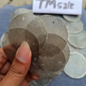 Puede incluir: Una mano sostiene un grupo de lentejuelas redondas, translúcidas y de color grisáceo, con pequeños agujeros. Las lentejuelas están superpuestas, creando un efecto texturizado. Una etiqueta blanca con el texto "TM sale" es visible en el fondo.