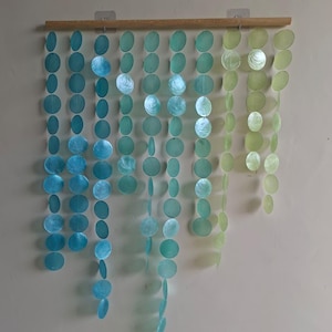 May include: A wall hanging featuring multiple vertical strands of circular, translucent shell discs suspended from a horizontal wooden rod. The discs transition in color from deep blue on the left to light green on the right, creating a gradient effect.