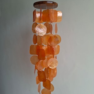 May include: An orange shell wind chime with a dark brown wooden top. The chime features various shapes, including circles and squares, all in shades of orange. The chime hangs from a thin string, creating a decorative and natural aesthetic.