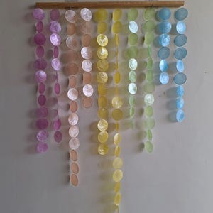 Peut inclure: Une suspension murale composée de multiples brins verticaux de disques de coquillages circulaires, translucides et irisés, suspendus à une tige en bois horizontale. Les disques sont disposés en dégradé de violet, pêche, jaune, vert et bleu.