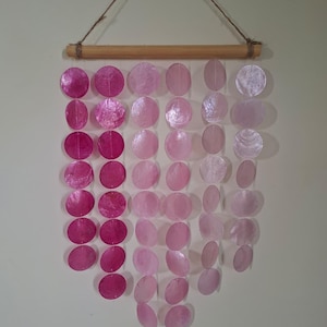 May include: A wall hanging featuring six vertical strands of circular, translucent capiz shells suspended from a horizontal wooden dowel. The shells transition in colour from deep magenta on the left to pale, iridescent white on the right.