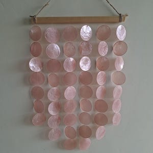 May include: A wall hanging featuring six vertical strands of circular, translucent, pale pink capiz shell discs suspended from a horizontal wooden dowel. A thin, natural-colored twine cord is attached to the ends of the dowel for hanging.