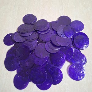 May include: A collection of circular purple sequins, each featuring two small holes for attachment. These flat, round embellishments have a glossy, iridescent finish and are scattered in a loose pile, showcasing their vibrant violet hue.