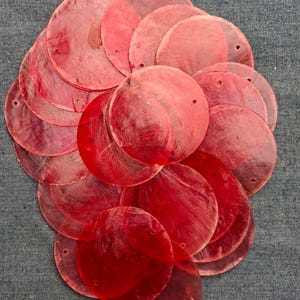 Könnte beinhalten: Eine Ansammlung von durchscheinenden, runden, roten Muschelscheiben mit kleinen Löchern, kreisförmig angeordnet. Die Scheiben haben eine leicht irisierende Qualität, was auf ein natürliches Material hindeutet. Der Hintergrund ist ein strukturiertes, blau-graues Gewebe.