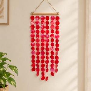 May include: A wall hanging featuring multiple vertical strands of circular, vibrant red shell discs suspended from a horizontal wooden rod. The strands are attached to the rod with a jute twine hanger, creating a cascading, layered display.