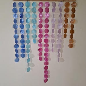 Puede incluir: Un colgante de pared con múltiples hilos verticales de discos circulares de concha nácar iridiscente suspendidos de una varilla de madera horizontal. Los discos están dispuestos en un degradado de color que va del azul al turquesa, rosa, morado y ámbar.