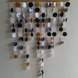 Peut inclure: Une décoration murale composée de multiples brins verticaux de disques circulaires dans des tons noirs, blancs, translucides et ambrés, suspendus à une tige en bois horizontale. Les disques sont disposés en longueurs variables.