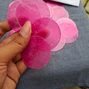 Pode incluir: Uma coleção de lantejoulas circulares rosa choque translúcidas. As lantejoulas são seguradas em uma mão e estão espalhadas sobre um tecido cinza. Um pequeno papel branco com o texto "TM sale" é visível no fundo.