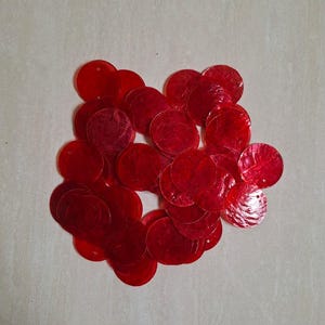Könnte beinhalten: Eine Ansammlung kreisförmiger roter Pailletten mit einer durchscheinenden, irisierenden Oberfläche. Jede Paillette hat ein kleines Loch nahe dem Rand zur Befestigung. Ein Teil eines Maßbandes ist in der Ecke sichtbar und zeigt Markierungen in Zentimetern.