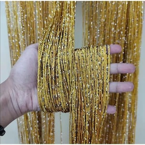 Puede incluir: Primer plano de un haz de hilo dorado con motas plateadas. El hilo se sostiene en una mano, mostrando su textura y la forma en que cuelga. El fondo está lleno del mismo hilo, creando un efecto brillante.